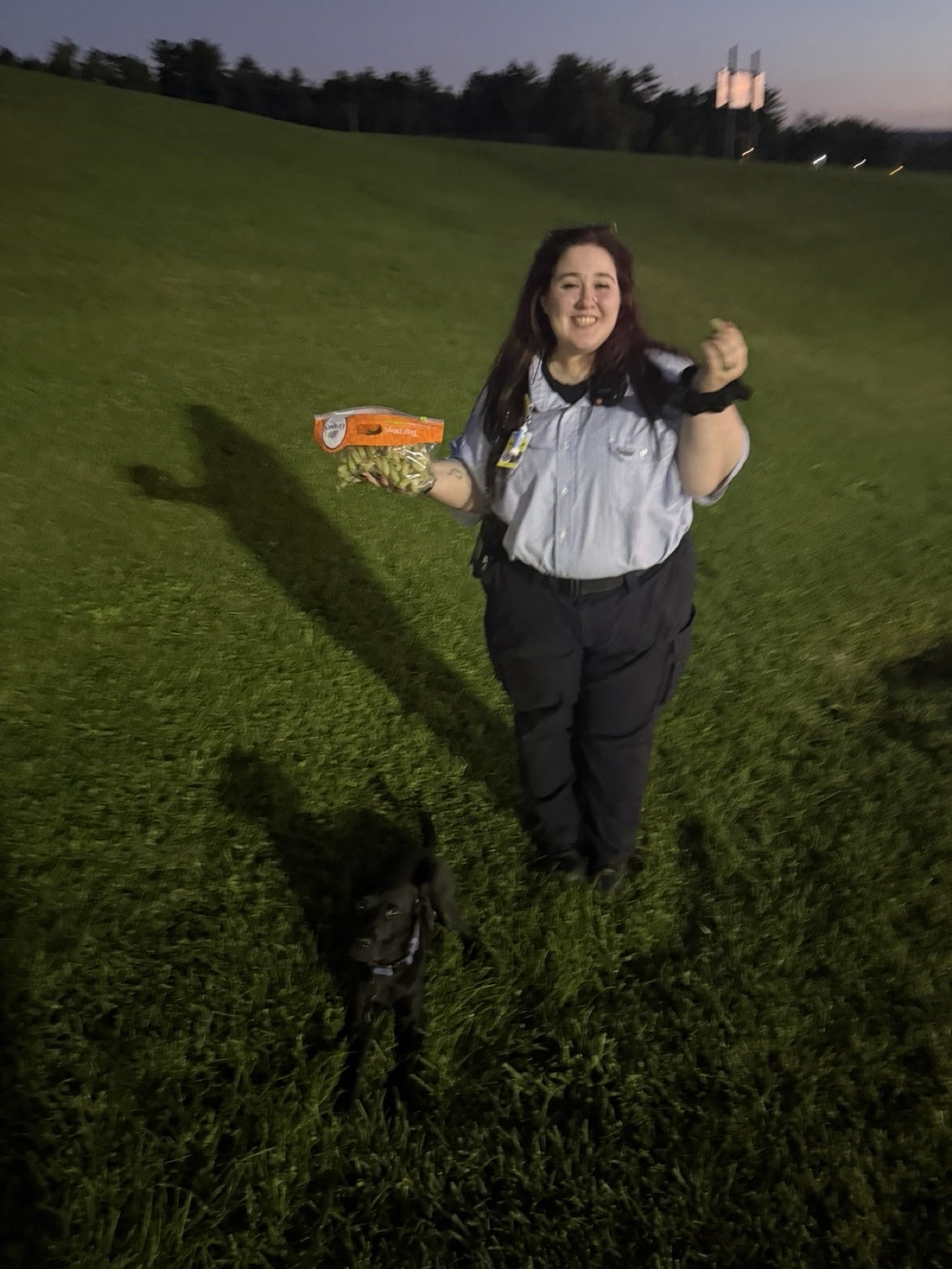 Savannah smiling on a grassy hill at dusk, holding a bag of snacks, with a black dog at her feet