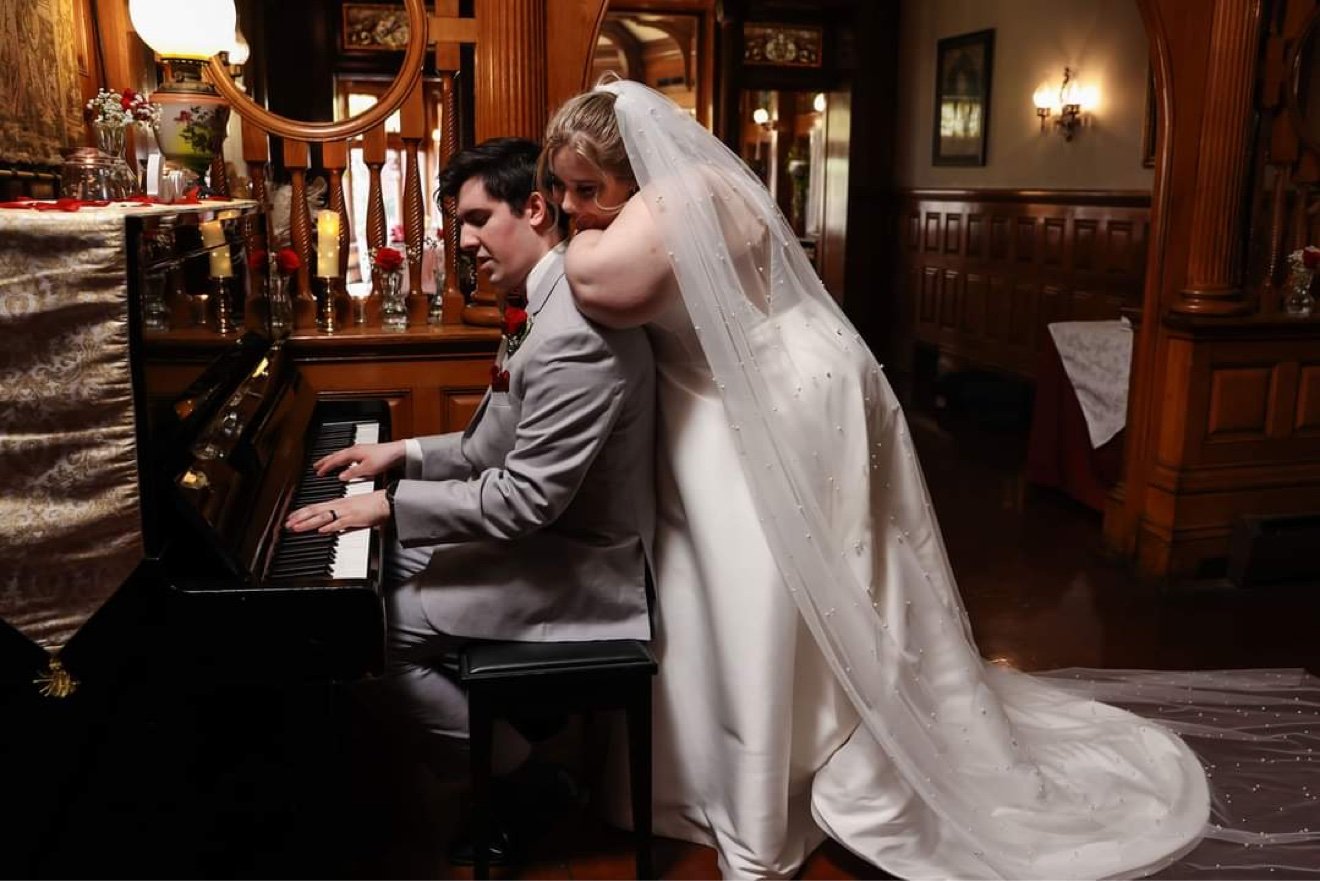 Axel playing the piano at their wedding while Savannah, in her veil, hugs him from behind
