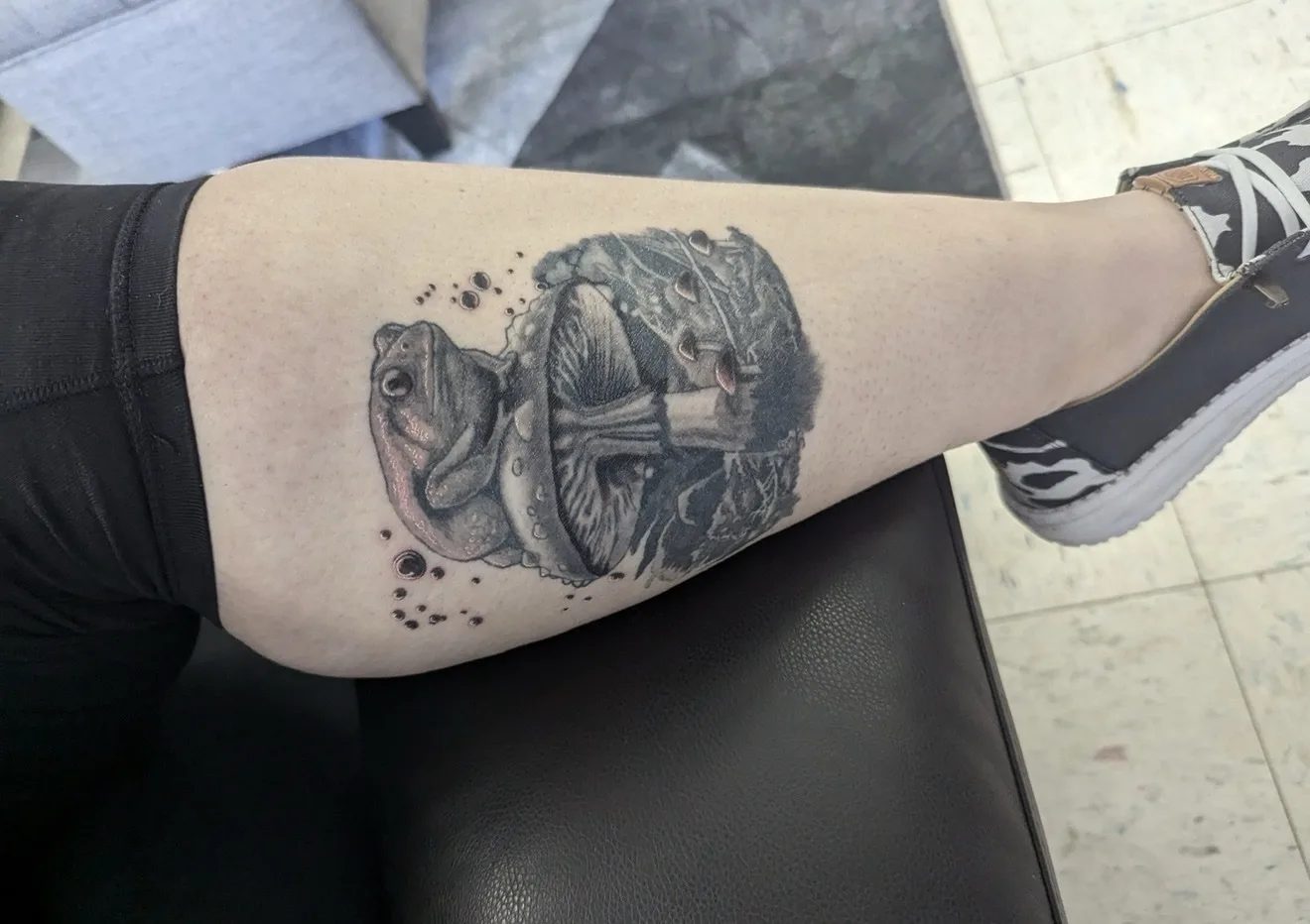 Black and grey tattoo of a frog sitting on a mushroom on a calf